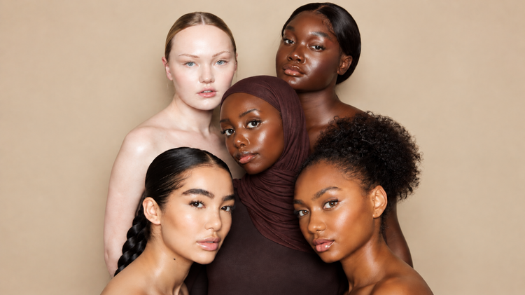Five women with radiant, glowing skin across a range of skin tones, posed together against a warm beige backdrop